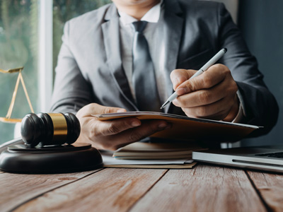 Ein Anwalt, der einen Stift benutzt, während er Dokumente überprüft. Im Vordergrund ein Richthammer und im Hintergrund ein Laptop.